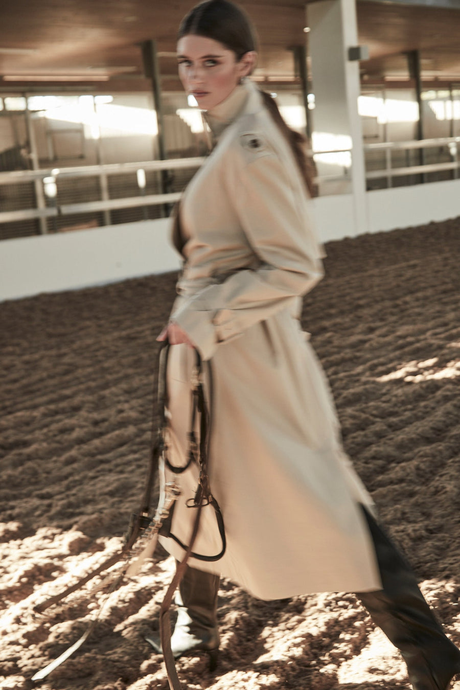 The E.L.S Celeste trench coat in beige, photographed in motion in a sunlit riding arena.