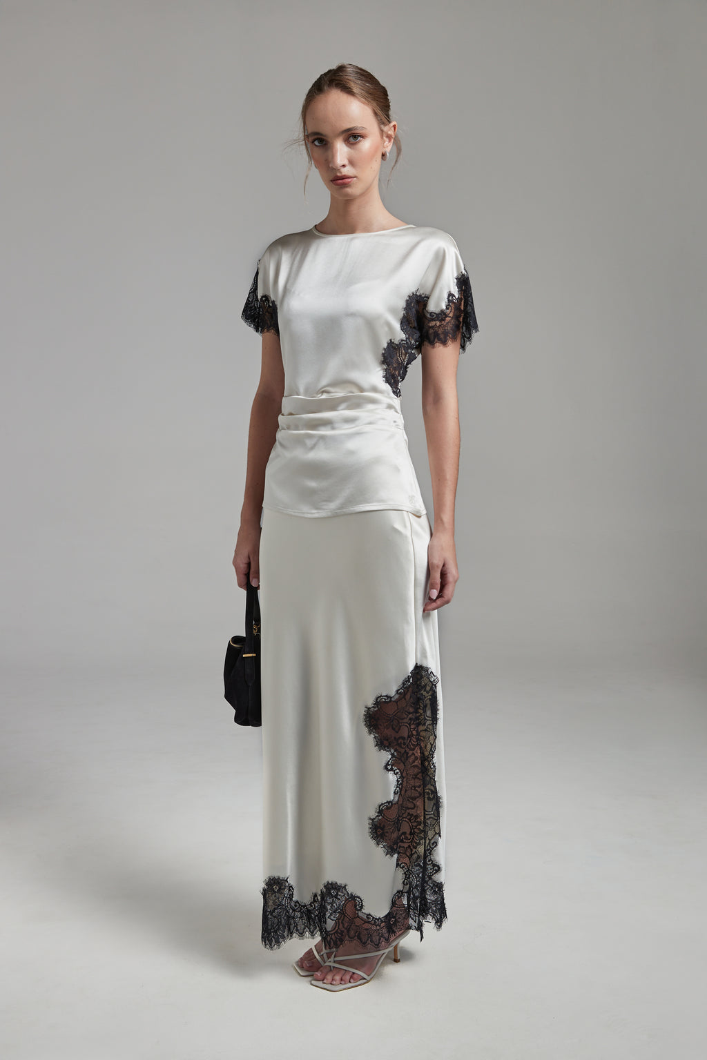 The Anouk Maxi Skirt in Feather White shown full-length from the front, featuring a satin finish, black eyelash-lace trim and a fluid ankle-length silhouette.