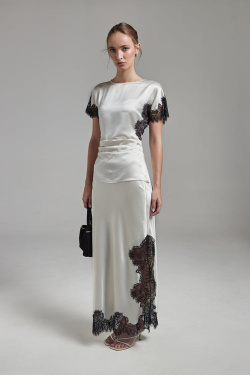 The Anouk Maxi Skirt in Feather White shown full-length from the front, featuring a satin finish, black eyelash-lace trim and a fluid ankle-length silhouette.