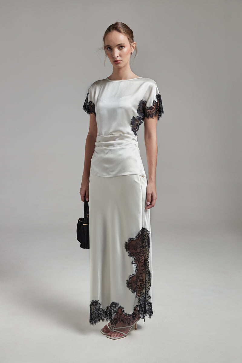 The Anouk Maxi Skirt in Feather White shown full-length from the front, featuring a satin finish, black eyelash-lace trim and a fluid ankle-length silhouette.