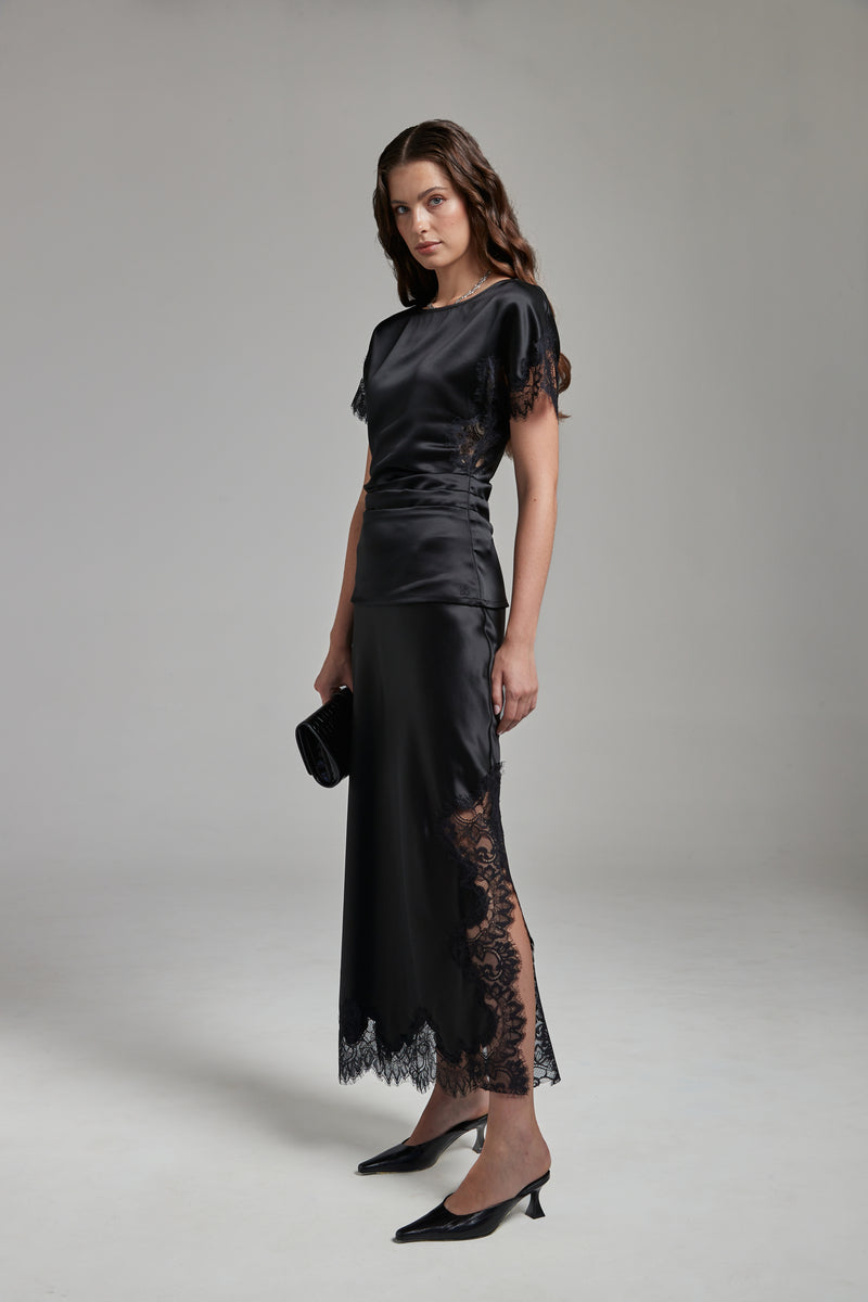 The Anouk Maxi Skirt in Midnight Black, a satin acetate lace skirt shown full-length from the side with tonal lace trim and ankle-length silhouette.