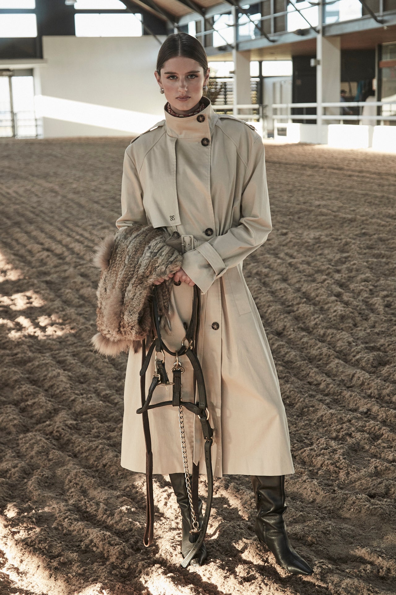 The Celeste Trench Coat in White Pepper, a beige cotton trench coat shown in a campaign image with structured silhouette, horse bridle and equestrian setting.