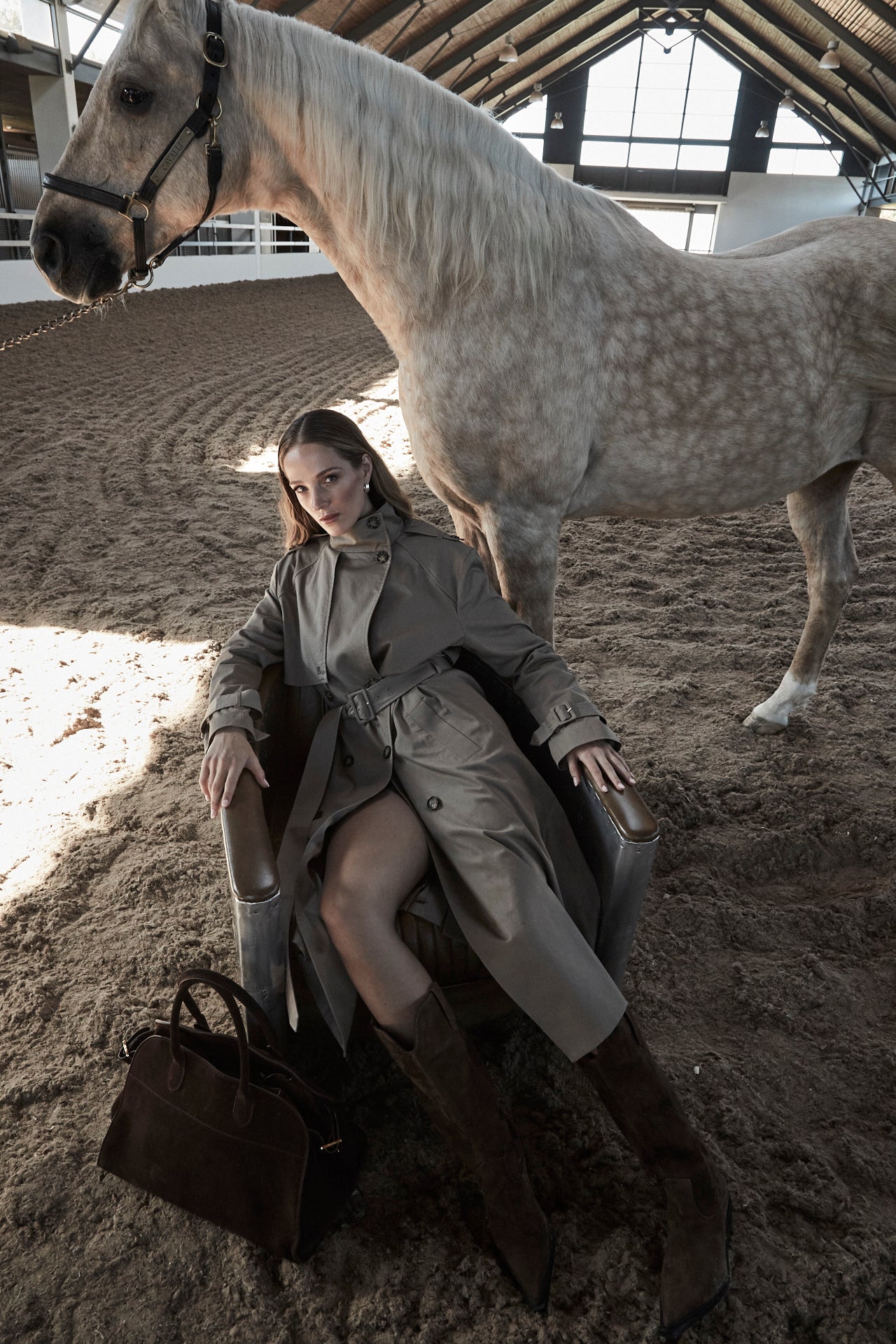 The Celeste Trench Coat in Desert Taupe, a cotton trench coat shown in an outdoor campaign image with structured silhouette and equestrian setting.