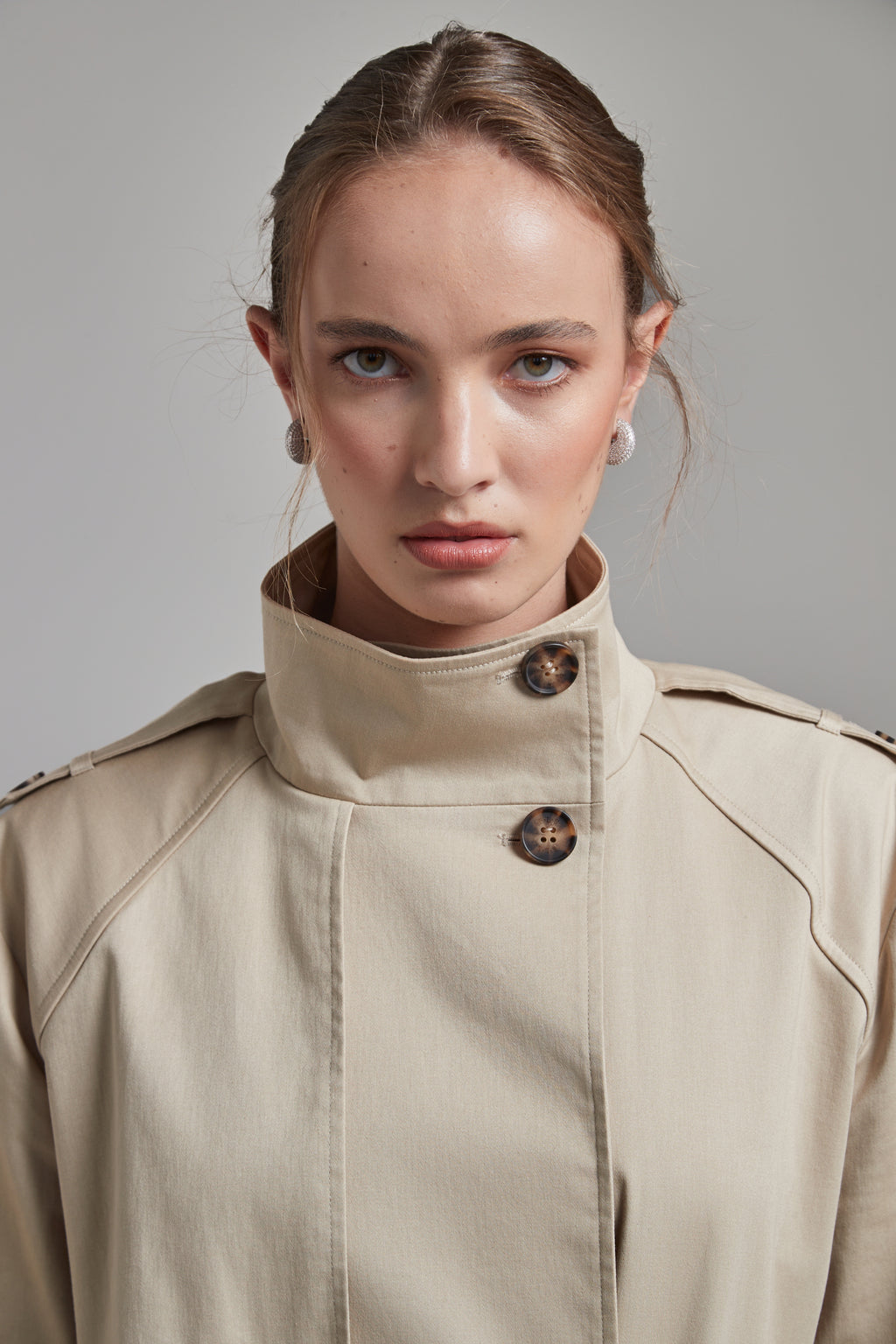 The Celeste Trench Coat in White Pepper, a beige cotton funnel neck trench coat shown front-on with structured silhouette and collar detail.