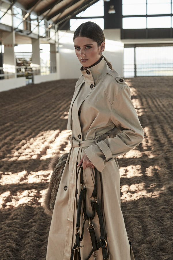 The Celeste Trench Coat in White Pepper, a cotton trench coat shown in a campaign image with structured silhouette, horse bridle and equestrian setting.