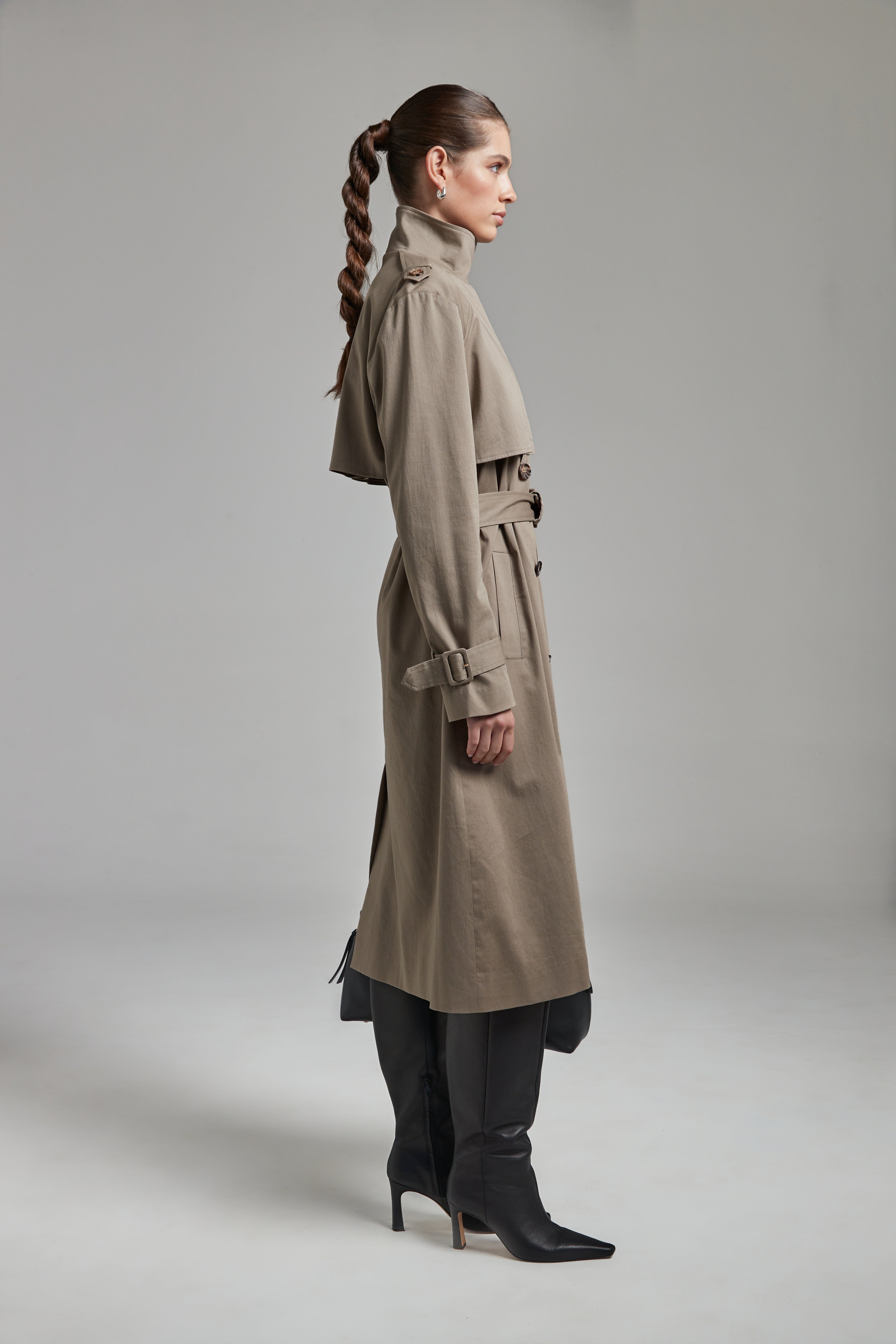 The Celeste Trench Coat in Desert Taupe shown full-length from the side with a funnel neckline and structured outerwear silhouette.