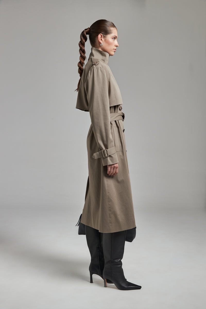 The Celeste Trench Coat in Desert Taupe shown full-length from the side with a funnel neckline and structured outerwear silhouette.
