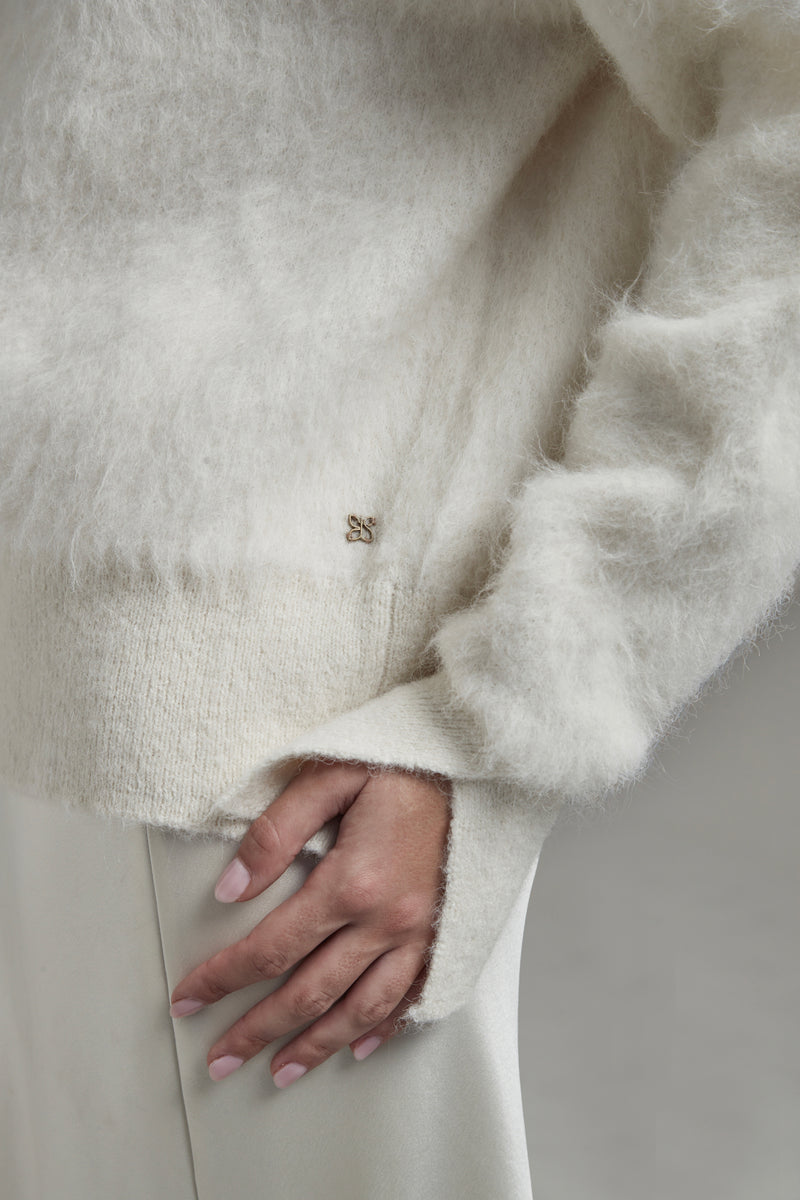 The Lauren Turtleneck in Feather White shown close-up with metal logo hardware detail and a brushed alpaca-blend texture.