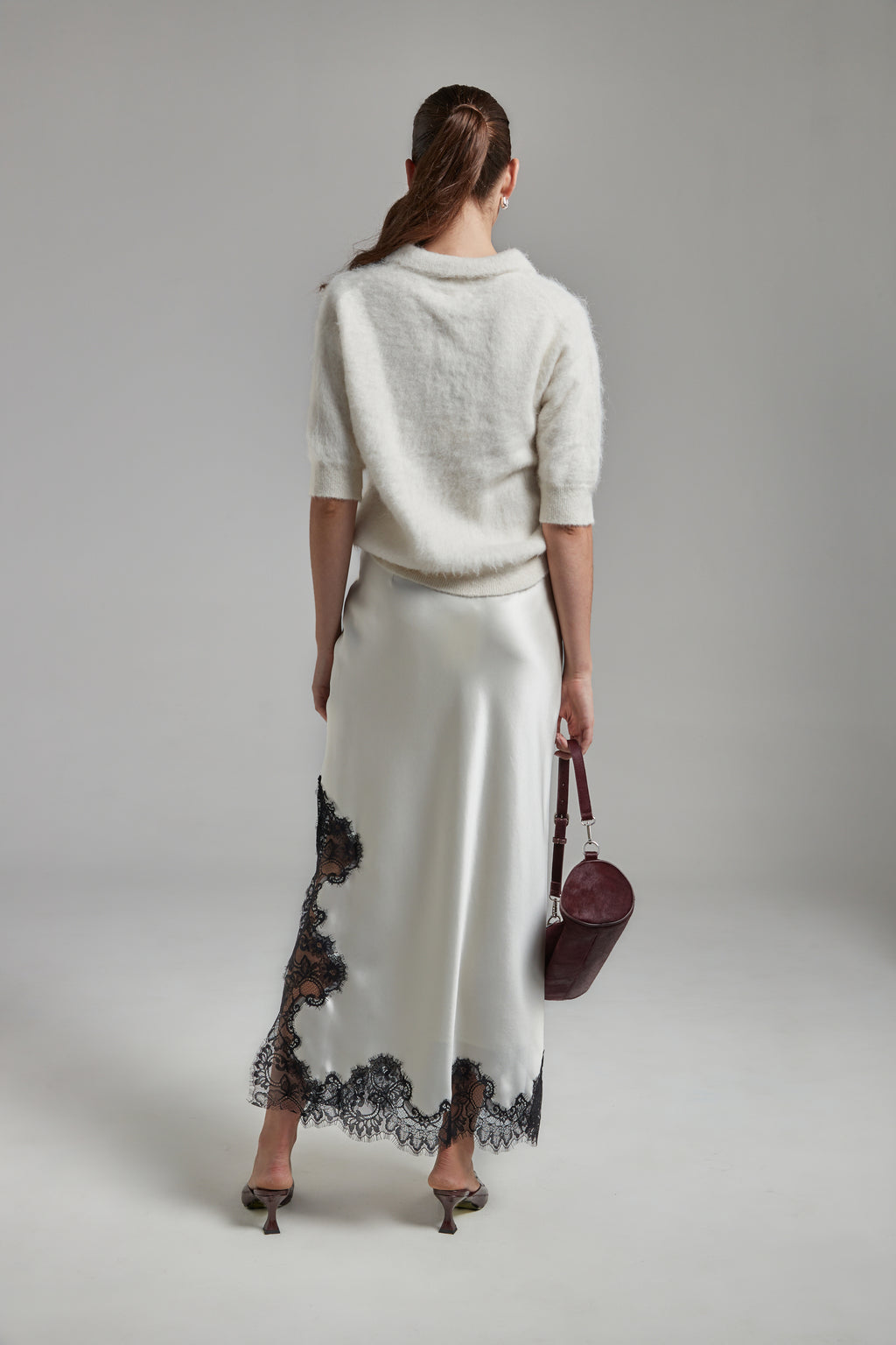 Model wearing the Stella Polo Knit and Anouk Maxi Skirt in Feather White, shown full-length from the back with a brushed alpaca-blend knit and black lace trim detailing.